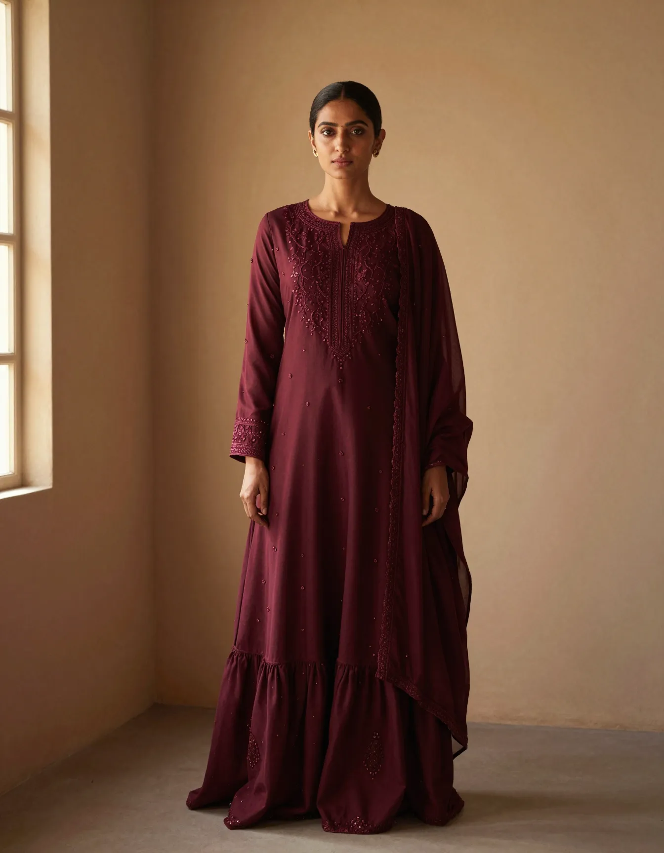 A model in a deep oxblood hand-embroidered sharara set against a plain plaster wall