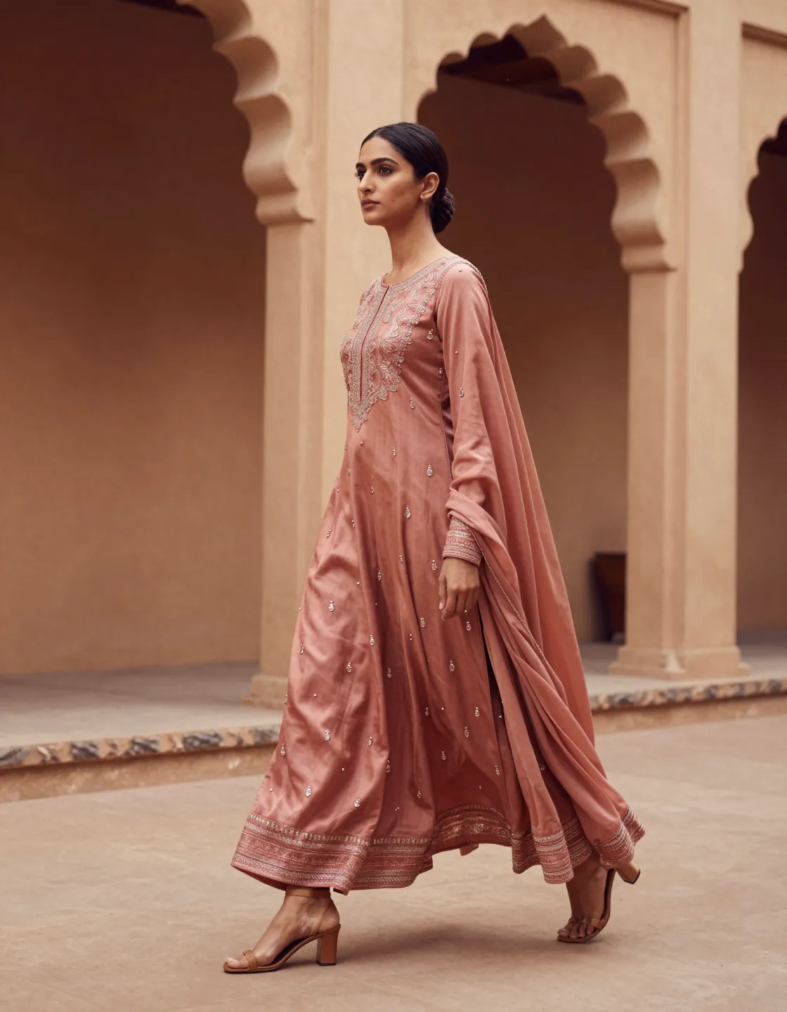 A model in a dusty rose anarkali walking through a warm plaster colonnade
